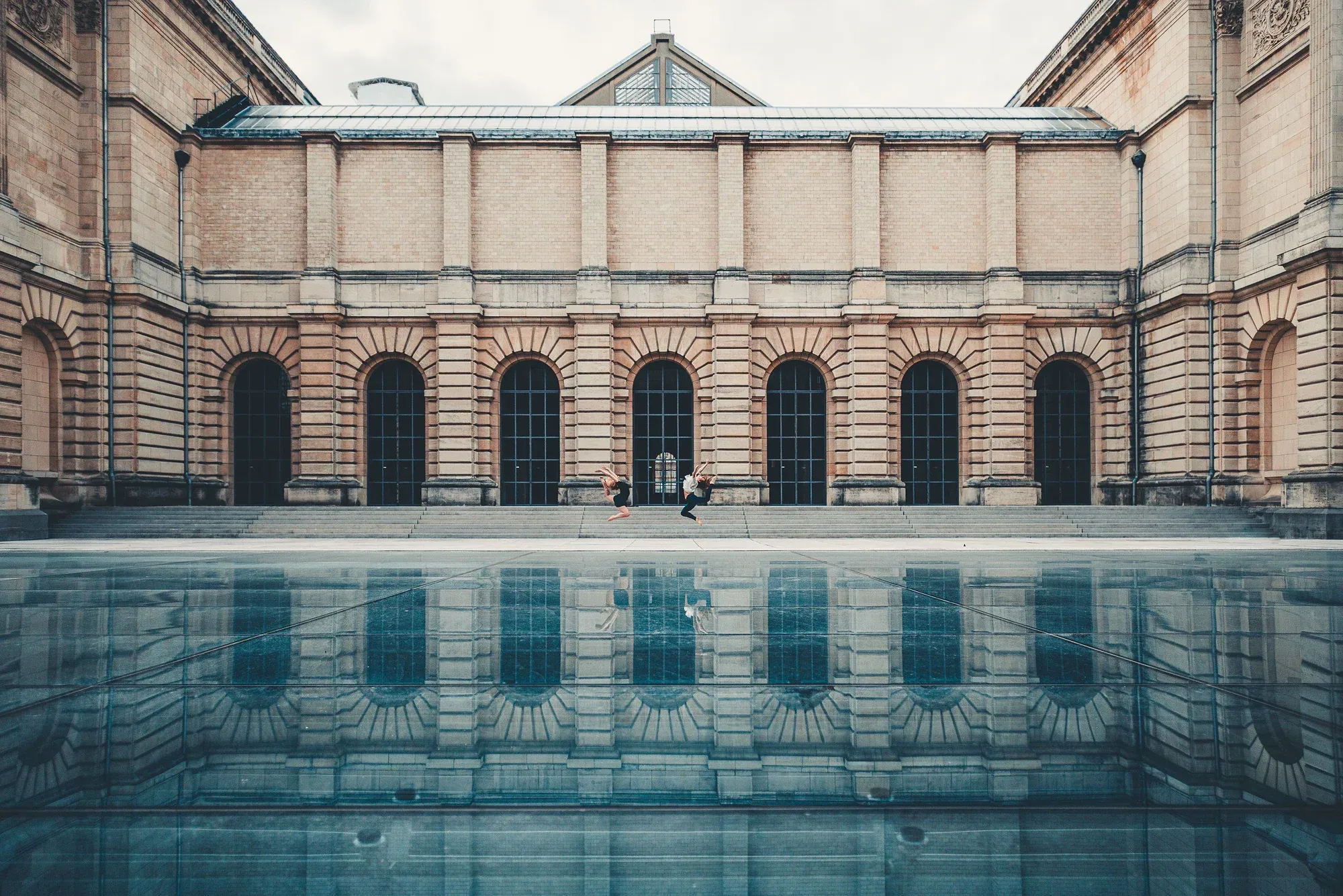 Architecture classique, reflet d'eau calme et silhouette de danseurs dans un vaste espace minéral.